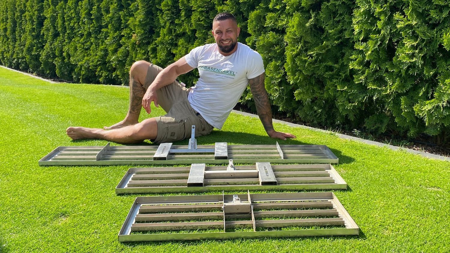 Drei RISISANI Rasenrakel in verschiedenen Breiten auf einer Rasenfläche, daneben sitzt ein Mann im RASENRAKEL-Shirt.